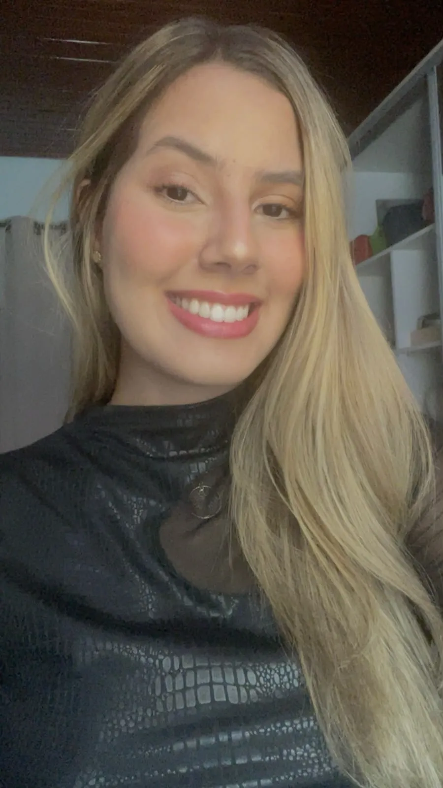 Letycia, de cabelos longos loiros e blusa preta, sorrindo em uma selfie.