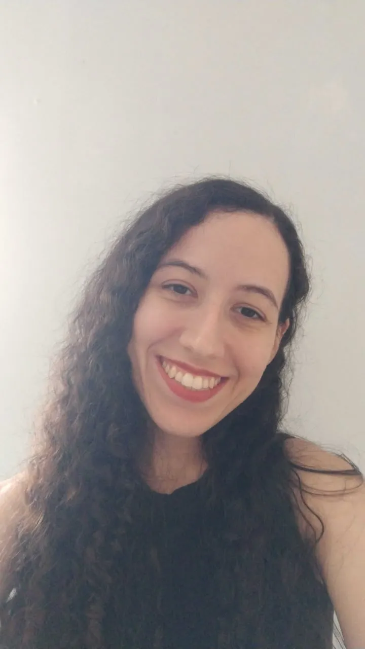 Mylena, de cabelos longos cacheados, sorrindo em frente a uma parede clara.