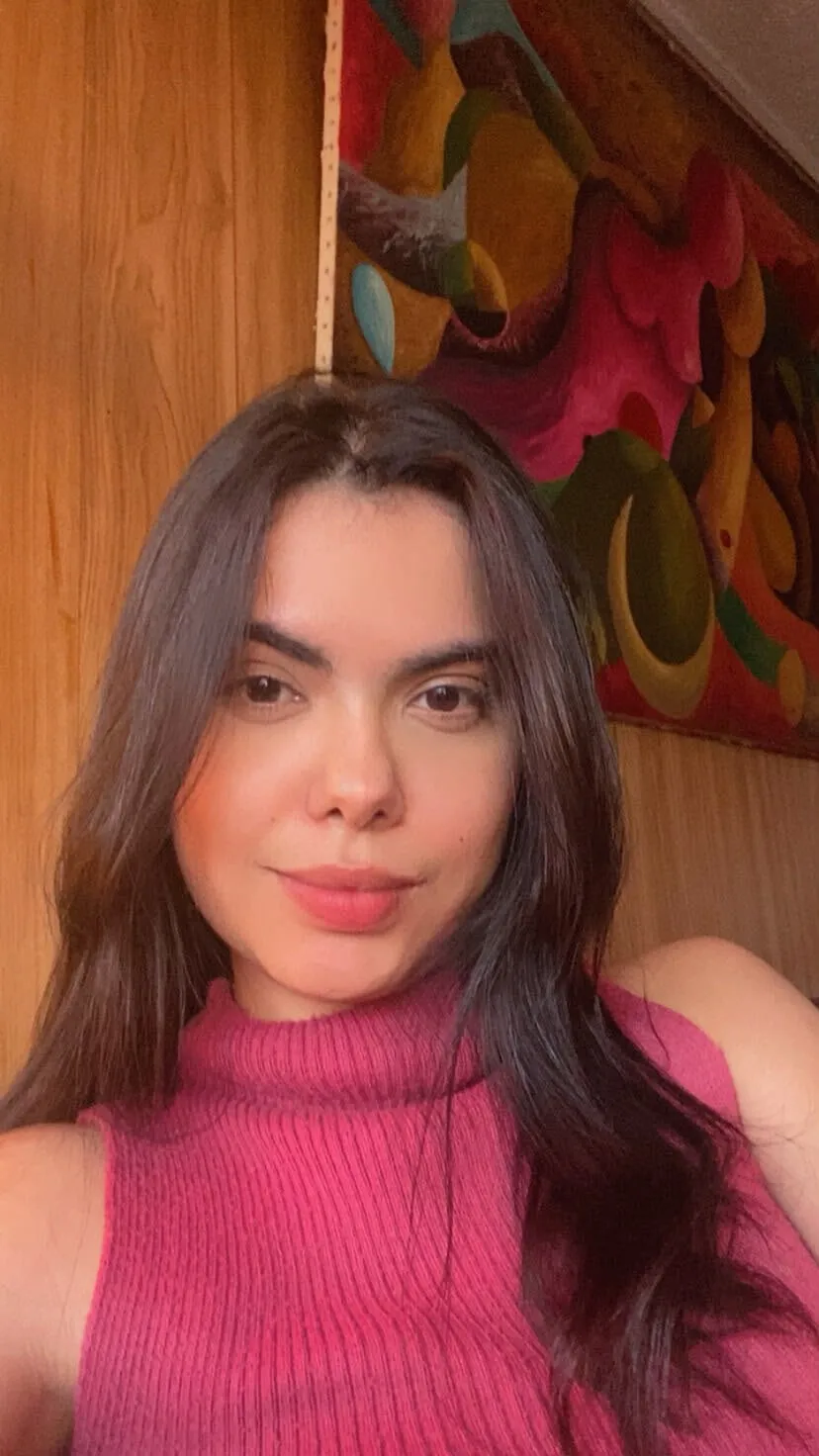 Paula, de cabelos longos escuros e blusa rosa, sorrindo diante de uma parede de madeira com um quadro colorido.